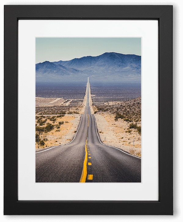 A long, straight desert highway cuts through arid land toward distant blue mountains, centered by a yellow line. Sparse vegetation and rocky hills frame the road, emphasizing vast space, isolation, and a sense of endless forward motion.
